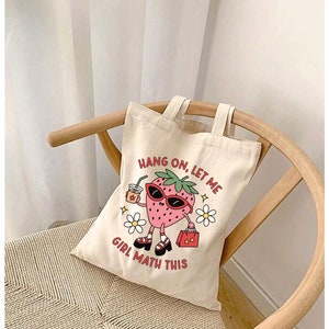 May include: A beige tote bag with a strawberry graphic and the text "Hang on, let me girl math this".