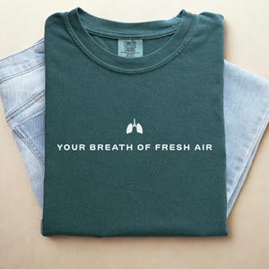 May include: A dark green t-shirt with white text that reads "YOUR BREATH OF FRESH AIR" and a white outline of lungs above the text. The t-shirt is folded and laid on a light brown surface.