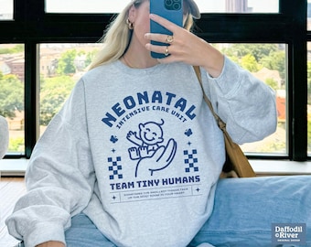 Op de afbeelding: Een grijs sweatshirt met een blauw grafisch ontwerp met de tekst "Neonatal Intensive Care Unit" en "Team Tiny Humans". De grafiek bevat ook een cartoonbaby in een blauwe romper.