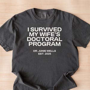 May include: A dark gray t-shirt with white text that reads "I survived my wife's doctoral program Dr. Junie Wells Est. 2025".