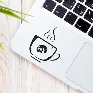 May include: A white laptop computer with a black keyboard and a black and white sticker of a coffee cup with steam coming from it on the top of the laptop.