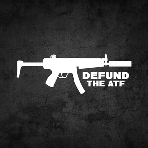 May include: A white silhouette of a submachine gun with the text "DEFUND THE ATF" below it. The image is on a dark gray textured background.