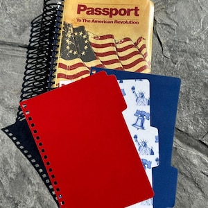 May include: A spiral-bound notebook with a cover featuring an American flag design and the text "Passport To The American Revolution." A red folder and a blue folder with a white and blue patterned insert are also visible.