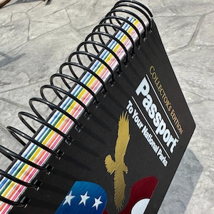 May include: Black spiral-bound "Collector's Edition Passport To Your National Parks" notebook. The cover features a gold eagle graphic and a stylized American flag design. The pages have colorful edges.