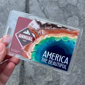 May include: Clear plastic holder containing an "America the Beautiful" annual pass. The pass features a colorful landscape design with the words "America the Beautiful" in white text. The pass also has the words "Annual Pass" and a mountain logo.