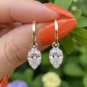 May include: A pair of gold hoop earrings, each featuring a pear-shaped, clear gemstone pendant. The earrings are held up against a blurred green and pink floral background.