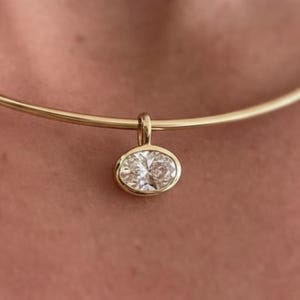 May include: A gold choker necklace featuring an oval-cut diamond pendant. The pendant is set in a gold bezel. The necklace is worn around the neck.