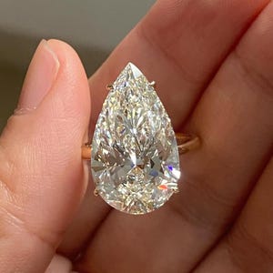 以下が含まれることがあります： ゴールドのバンドにペアシェイプのダイヤモンドが付いた指輪。透明でファセットカットされたダイヤモンドが光を捉えています。指輪は手で持たれており、宝石のサイズと輝きを際立たせています。ジュエリーです。