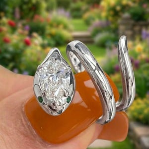 以下が含まれることがあります： 洋梨型のダイヤモンドの頭と2つの小さな緑色の宝石を眼に使用した、銀色のスネークリング。 リングは指に巻き付き、ユニークでエレガントなデザインを作り出しています。 オレンジ色のマニキュアを塗った手でリングが示されています。