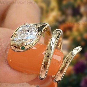 May include: A gold-colored ring in the shape of a snake, with a large, clear, pear-shaped diamond as the head. The snake's head has small green accents. The ring wraps around the finger in a spiral design. The text "Caratline Diamonds" is visible.