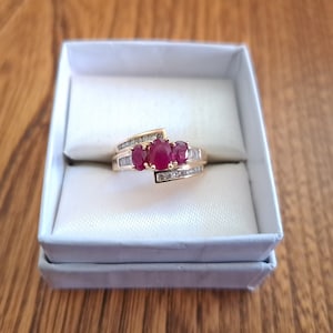 Peut inclure: Une bague en or avec trois pierres précieuses rouges de forme ovale et de plus petites pierres transparentes. La bague est présentée dans une boîte blanche sur une surface en bois. Le design de la bague présente un motif tourbillonnant.