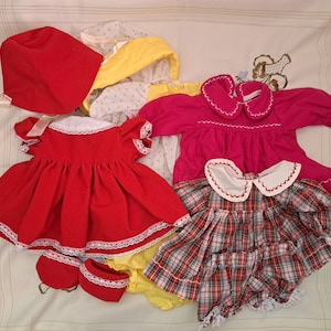 May include: A selection of vintage baby clothes in various colours. Includes a red corduroy dress with white lace trim, a pink dress, a plaid dress, bonnets, and matching shoes. The dresses have white collars.