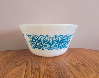 May include: White mixing bowl with a teal floral design. The bowl has a rounded shape and a wide rim. The teal design features stylized flowers and leaves. The bowl is sitting on a wooden surface.