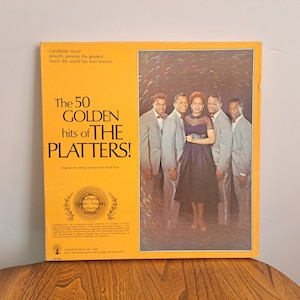 Peut inclure: Une pochette d'album de disque jaune avec une photo d'un groupe vocal, The Platters, sur le devant. Le titre de l'album est "The 50 Golden Hits of The Platters!" La pochette de l'album comprend également le texte "Candlelite Music proudly presents the greatest music the world has ever known."