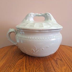 May include: A white ceramic tureen with a lid featuring two bird figures. The tureen has a handle and decorative floral patterns. The lid has a geometric design. The tureen is sitting on a wooden table.