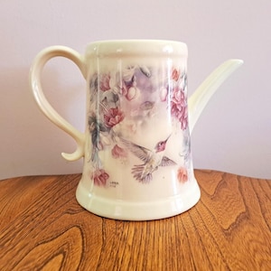 May include: Cream ceramic watering can with a curved handle and spout. The can features a floral and hummingbird design in pink, purple, and grey. The base is slightly flared. Measurements are in centimetres.