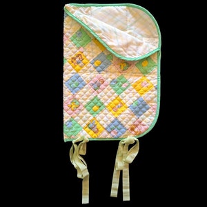May include: A quilted baby bib in pastel shades. The bib has a diamond pattern with cartoon characters in yellow, blue, pink, and green squares. It has a green trim and ties.