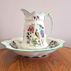 May include: A white porcelain jug and bowl set with a green trim and floral pattern. The jug has a handle and a bird design. The bowl is round and has a scalloped edge.