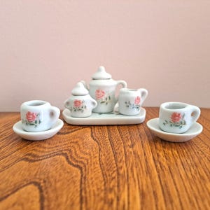 May include: A miniature porcelain tea set with a floral rose design. The set includes a teapot, sugar bowl, creamer, and two teacups with saucers. The pieces are white with pink and green accents.