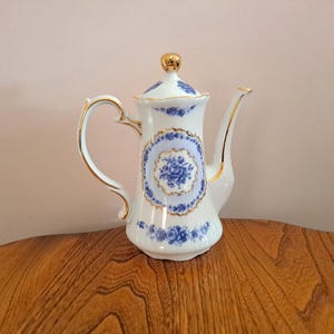 May include: A white porcelain teapot with a blue and gold floral design. The teapot has a gold lid and a gold handle.