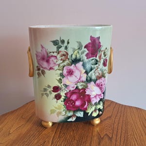 May include: A rectangular vase with a floral design featuring pink and burgundy roses, green leaves, and gold accents. The vase has gold handles and small gold feet. The background is a light green with a gradient effect.