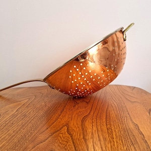May include: A copper-colored colander with a long handle and small perforations. The colander has a shiny, reflective surface and a brass-colored rim and handle. The kitchen utensil is angled on a wooden surface.