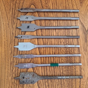 May include: An assortment of metal spade drill bits, arranged on a wooden surface. The bits vary in size and shape, with flat, paddle-like heads and cylindrical shafts. Some bits have markings, and one has a green band.