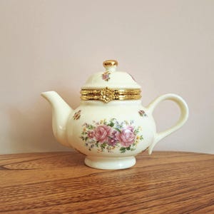 May include: Cream-coloured ceramic teapot with a floral design of pink roses and green leaves. The lid and rim are accented with gold-coloured metal. The teapot has a small handle and spout.