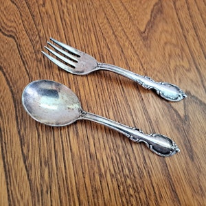May include: A silver fork and spoon set with decorative handles. The fork has four tines, and the spoon has a rounded bowl. The utensils are placed on a wooden surface, reflecting light. The handles feature an ornate pattern. The set is likely for dining.