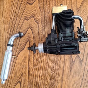 May include: A model engine with a black body, silver exhaust pipe, and gold-colored top. The engine is designed for model aircraft or vehicles. The exhaust pipe is detached, showing its silver finish and black rings.