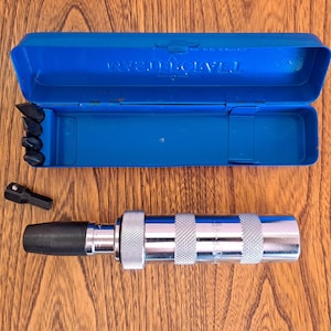 May include: A chrome impact driver with a black rubber tip and a blue plastic case. The case is open, revealing several black bits. The driver has a textured grip and is lying on a wooden surface.