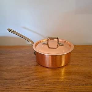 Peut inclure: Une casserole en cuivre poli avec un couvercle assorti et une longue poignée en laiton robuste. La casserole ronde présente une surface métallique lisse et réfléchissante ainsi qu'un couvercle sécurisé avec une poignée centrale en laiton.