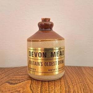 May include: A vintage ceramic bottle of Devon Mead, Britain's oldest drink, with a cork stopper. The bottle has a brown top and a beige body with a gold label. The label reads "Devon Mead" and "Britain's Oldest Drink".