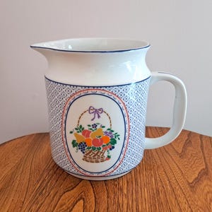 May include: White ceramic jug with a blue and white patterned design. The jug features a basket of fruit with a purple bow.