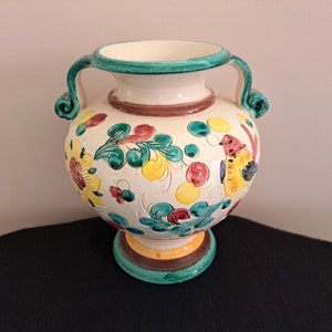 May include: A white ceramic vase with green, yellow, red and brown floral and bird designs. The vase has two handles and a round base.