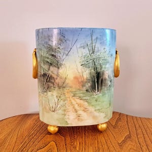 May include: A cylindrical decorative vase with a hand-painted landscape scene. The vase features a path through a forest, with gold-coloured handles and feet. The colour palette includes blues, greens, and yellows.