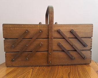 May include: A vintage wooden sewing box with a handle. The box is made of light brown wood and features a tiered design with angled wooden supports. The handle is arched and made of the same wood. The box is designed to hold sewing supplies.
