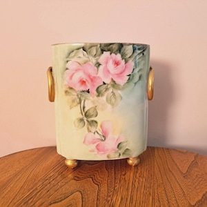 May include: A vintage porcelain vase with gold accents and a floral design featuring pink roses and green leaves. The vase has two gold handles and sits on four gold feet.