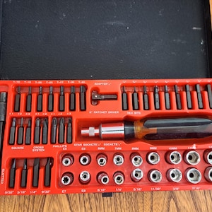 May include: A red toolbox containing a comprehensive set of tools, including screwdriver bits, sockets, and an extension bar. The set features slotted, square, Phillips, and hex bits, plus a ratchet driver. The toolbox is open, displaying the organised tools.