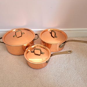 Peut inclure: Ensemble de trois casseroles en cuivre avec couvercles et poignées en laiton. Les casseroles sont de tailles différentes, la plus grande ayant deux poignées et les plus petites une longue poignée. Le cuivre est brillant.