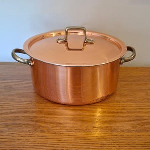 May include: A round copper cooking pot with a matching lid. The pot features two curved brass side handles and a central brass handle on the lid. The exterior surface has a polished metallic finish with a warm reddish-brown copper tone.