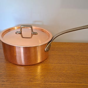 Peut inclure: Une casserole en cuivre poli avec un couvercle assorti et une longue poignée métallique incurvée. La casserole présente une surface extérieure lisse et réfléchissante, ainsi qu'une poignée robuste fixée par deux rivets visibles.