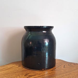 May include: A dark blue, antique stoneware crock with a wide mouth and a slightly rounded body. The crock has a glossy finish and sits on a wooden surface. This vintage kitchenware piece is likely used for food storage.
