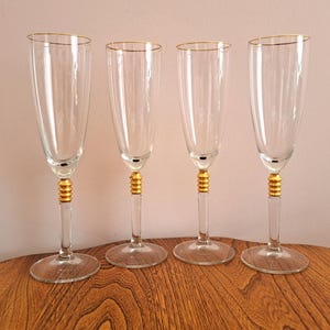 May include: Four clear glass champagne flutes with gold rims and a gold band around the stem. The flutes are arranged in a row on a wooden surface.