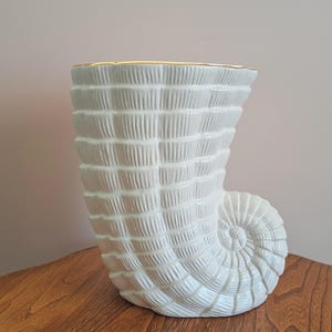 Peut inclure: Un vase en céramique blanche en forme de coquille de conque avec une surface texturée et nervurée, ainsi qu'un bord fin peint en or. Le récipient présente une base en spirale et une large ouverture supérieure, imitant la forme naturelle d'un coquillage.