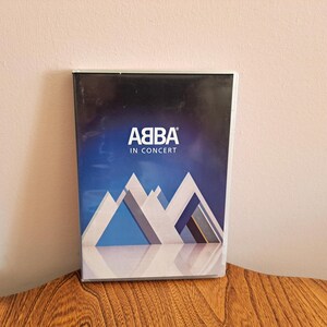 May include: DVD case for ABBA In Concert. The cover features the band's logo in white, with the words "IN CONCERT" below. The background is a gradient of blue and white, with a stylized mountain range in the foreground.