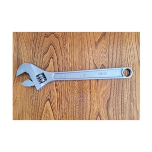May include: A silver adjustable spanner with the word "GRAY" printed on the handle. The spanner is lying on a wooden surface. The spanner has an adjustable jaw and a hole at the end of the handle.