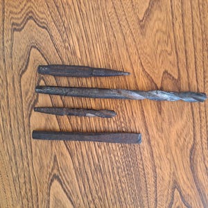 May include: A collection of vintage metal tools, including chisels and a drill bit, arranged on a wooden surface. The tools are dark in color, showing signs of wear and use. The arrangement suggests a focus on woodworking or metalworking.