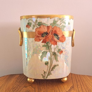 May include: A decorative, rectangular vase with a pearlescent finish, featuring a gold rim and handles. The vase is adorned with orange poppy flowers and green leaves. It stands on three gold feet.