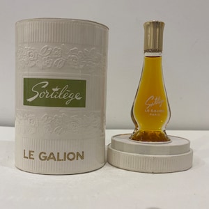 May include: A small bottle of Sortilège perfume by Le Galion Paris. The bottle is clear glass with a gold cap and a white label with the brand name and city. The bottle is sitting on a white lid with a white box behind it.